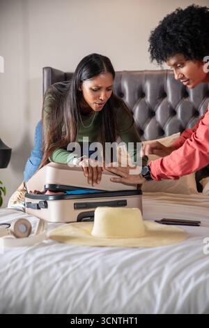 Couple diversifié penché sur le lit blanc appuyant sur le couvercle de valise à côté du chapeau de soleil de paille Banque D'Images