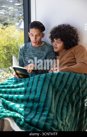 Divers amis masculins assis sur le siège de la fenêtre partageant une couverture verte lisant le livre de poche par la grande fenêtre Banque D'Images