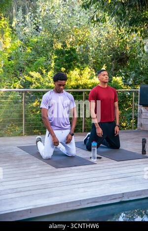 Divers amis masculins en vêtements de sport agenouillant sur des tapis de yoga sur une terrasse en bois par la piscine tenant smartphone Banque D'Images