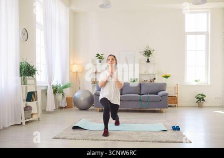 Femme senior portant des vêtements de sport debout dans le salon à la maison exerçant sur le tapis de yoga. Banque D'Images
