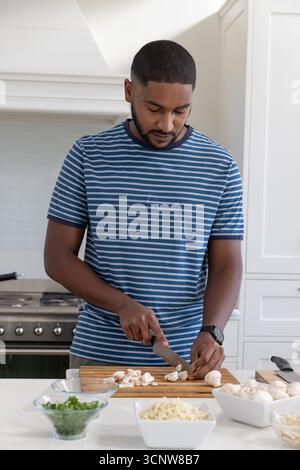 Homme afro-américain tranchant des champignons sur une planche à découper sur l'île à côté de bols d'herbes et de fromage Banque D'Images
