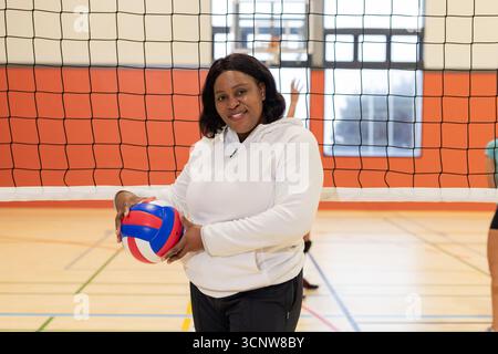 Femme afro-américaine mature tenant le ballon près du filet de volley-ball, face à la caméra dans la salle de sport Banque D'Images