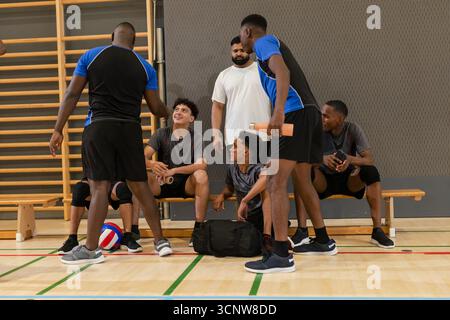 Divers coéquipiers masculins de volley-ball avec entraîneur se rassemblant sur le banc dans la salle de gym autour de l'équipement de volley-ball Banque D'Images