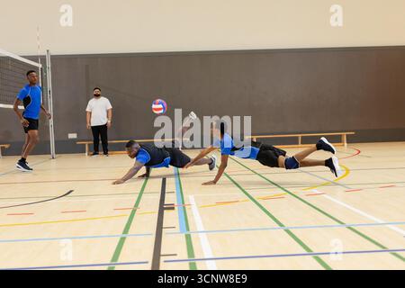 Divers coéquipiers masculins plongeant vers le volleyball en plein air et observant l'entraîneur sur le terrain près du filet Banque D'Images