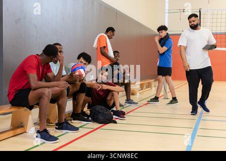 Équipe masculine diversifiée de volley-ball sur banc en bois tenant des balles de volley-ball dans la salle de gym pendant que l'entraîneur marche avec TAB Banque D'Images
