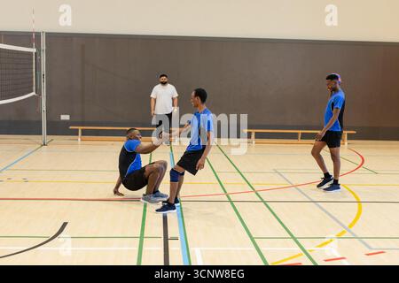 Équipe masculine diversifiée de volley-ball aidant son coéquipier sur le terrain de volley-ball avec genouillère et poteau de filet Banque D'Images