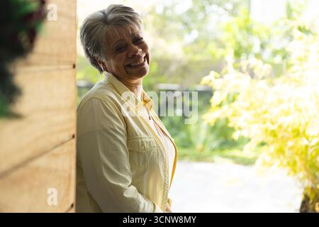 Femme senior appuyée contre le mur de porche en bois avec couronne face aux plantes de jardin souriant à la lumière du soleil Banque D'Images