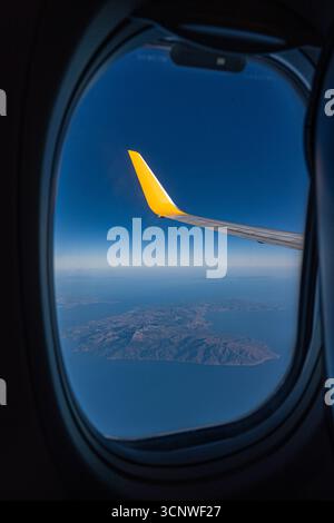 Une fenêtre d'avion révèle une vue captivante sur le ciel bleu et l'océan serein en contrebas, avec un bout d'aile jaune proéminent en vue, illustrant l'essen Banque D'Images