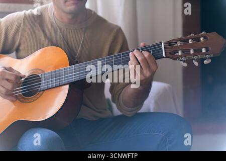 Un homme tape une guitare acoustique dans le confort de sa maison, portant une tenue décontractée. Un éclairage doux crée une atmosphère détendue et accueillante, parfaite fo Banque D'Images