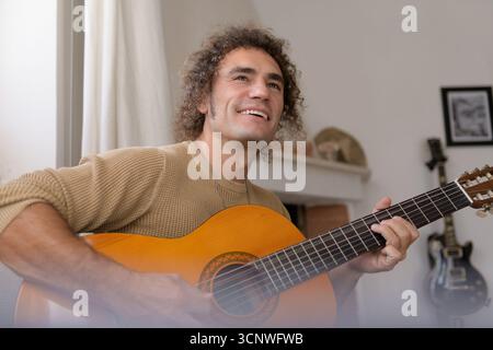 Un homme assis confortablement à la maison tout en tapant une guitare acoustique. La chambre est confortable, créant une atmosphère détendue parfaite pour profiter de la musique et l Banque D'Images
