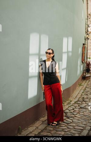Une femme à la mode se tient contre un mur éclairé par le soleil, vêtue d'une tenue élégante avec des lunettes de soleil, marchant dans une rue pavée pittoresque dans un pictu Banque D'Images