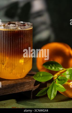 Un café rafraîchissant servi dans un verre transparent avec une base de jus d'orange éclatante et de la glace. Les oranges fraîches et les feuilles vertes ajoutent une touche de nature à la Banque D'Images