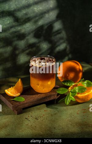 Un verre de café butin repose sur une plate-forme en bois, entourée de tranches d'orange vives et de feuilles vertes. La riche couleur du café contraste magnifiquement Banque D'Images