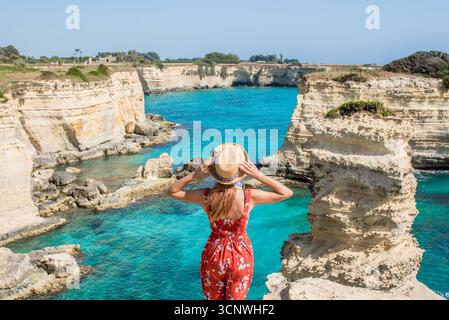 Une femme vêtue d'une robe d'été et d'un chapeau regarde les magnifiques eaux turquoises et les falaises spectaculaires de Torre Sant Andrea dans les Pouilles, en Italie, incarnant la sérénité Banque D'Images