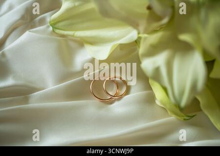 Deux anneaux de mariage dorés reposent élégamment sur un tissu doux et blanc entouré de délicats accents floraux, créant une atmosphère sereine et romantique parfaite Banque D'Images