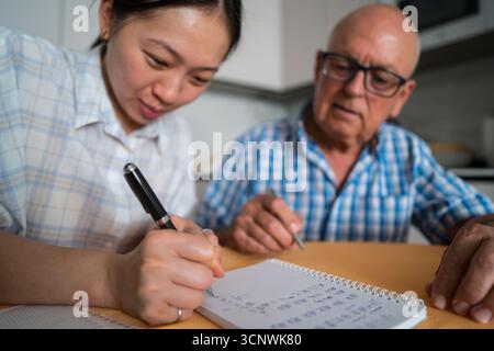 Un homme âgé est engagé dans l'apprentissage du mandarin, assisté par une enseignante. Ils se concentrent sur un cahier rempli de caractères chinois, mettant l'accent sur cro Banque D'Images