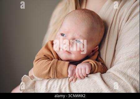 Un bébé serein est bercé avec amour dans les bras d'un soignant, portant une tenue brune confortable. L'expression du contenu du bébé et les yeux bleus reflètent le confort et Banque D'Images