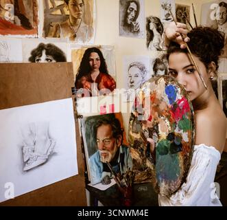 Une jeune femme peintre se tient confiante dans son home studio, entourée de ses œuvres d'art détaillées et expressives, tenant une palette colorée et un pinceau Banque D'Images
