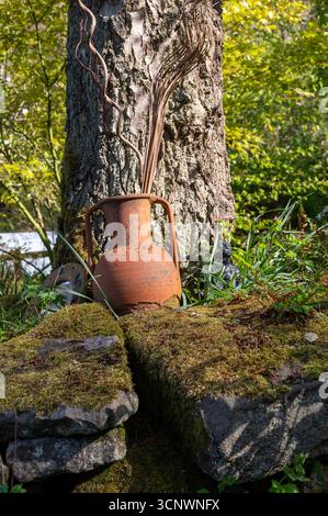 Amphore en terre cuite à la base d'un vieux bouleau avec des pierres couvertes de mousse, photographiée en avril dans un jardin boisé, Royaume-Uni Banque D'Images