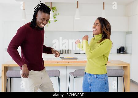 Couple diversifié dansant et riant dans la cuisine sous des lumières suspendues blanches près de l'île avec des pâtisseries Banque D'Images