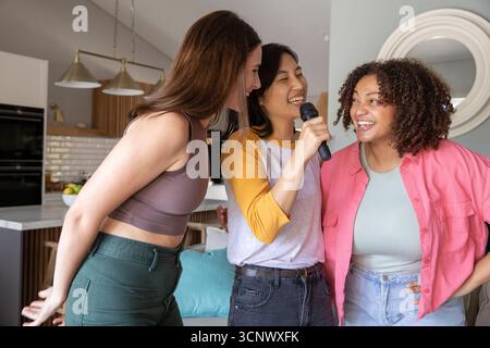 Diverses amies chantant karaoké dans le salon avec microphone à main et coussins sarcelle Banque D'Images