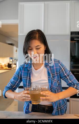 Femme asiatique debout derrière l'îlot de cuisine tenant trois tasses en verre avec du café près de la cafetière Banque D'Images