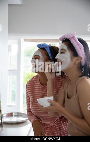 Divers amis féminins appliquant des masques faciaux à la vanité de salle de bains à la maison en utilisant bol en céramique et spatule Banque D'Images
