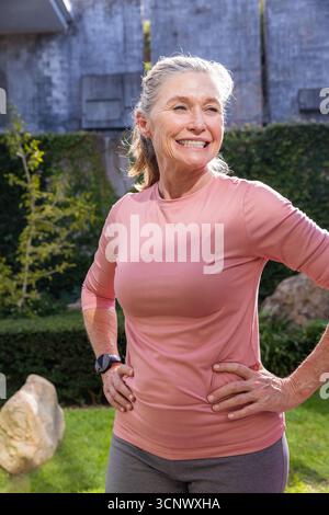 Femme âgée souriante debout dans l'arrière-cour portant un haut athlétique, smartwatch au milieu de la haie lierre rocheuse Banque D'Images