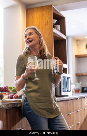 Femme blanche senior appuyée sur le comptoir en bois dans la cuisine tenant le verre à vin à côté de produire Banque D'Images