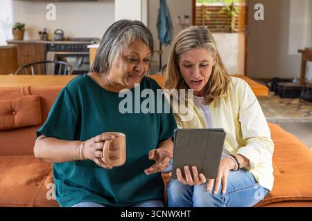 Divers amis féminins seniors assis sur le canapé dans le salon tenant la tasse à café et la tablette Banque D'Images