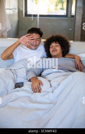 Divers partenaires masculins protégeant les yeux et câlins sous couette sur le lit dans la chambre avec smartphone Banque D'Images