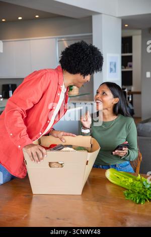 Couple diversifié déballer l'épicerie à la table à manger dans la cuisine avec boîte, céleri, téléphone Banque D'Images