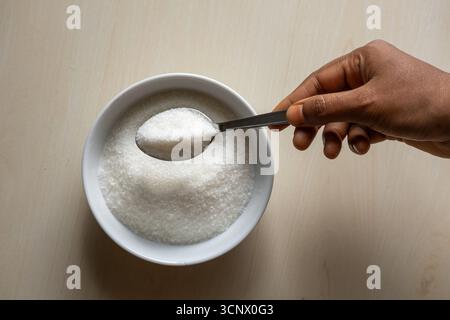 Un bol blanc est rempli de sucre sur fond de bois. Main de femme tenant une cuillère remplie de sucre. Ingrédients alimentaires pour la cuisson de bonbons ou desserts. Banque D'Images