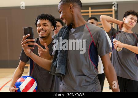Divers partenaires masculins de volleyball interagissant sur smartphone dans la salle de gym de l'école avec balle et serviette Banque D'Images