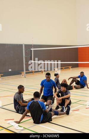 Divers coéquipiers masculins assis et parlant sur le sol de la salle de gym sous le filet de volley-ball avec une serviette blanche Banque D'Images
