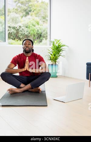 Homme afro-américain assis les jambes croisées sur le tapis de yoga à la maison, pressant les mains dans la prière près de l'ordinateur portable Banque D'Images