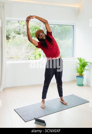 Homme afro-américain effectuant un étirement latéral sur un tapis de yoga dans une pièce minimale lumineuse avec tablette Banque D'Images