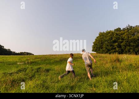 Un père et son fils profitent de leur temps à l'extérieur, courant ensemble à travers un champ vert luxuriant sous un ciel bleu clair. Le soleil brille brillamment comme eux Banque D'Images
