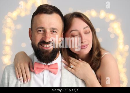Faire une proposition en mariage. Femme avec bague de fiançailles et son fiancé heureux embrassant sur fond gris clair avec des lumières floues, effet bokeh Banque D'Images