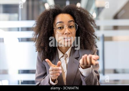 Portrait en gros plan d'une jeune femme afro-américaine en costume et lunettes dans un bureau, parlant lors d'un appel vidéo et comptant sur ses doigts jusqu'à la caméra. Banque D'Images