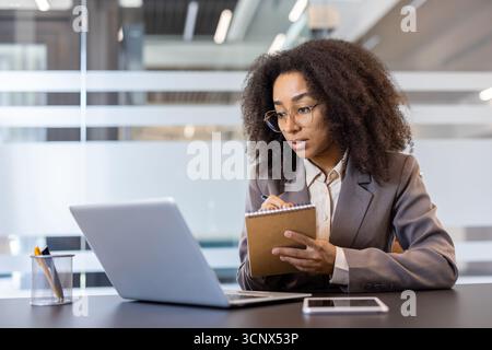 Concentrée jeune femme afro-américaine en costume d'affaires assis dans le bureau au bureau devant l'ordinateur portable et prenant des notes. Banque D'Images