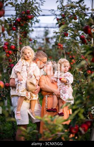 Une famille joyeuse passe une journée ensoleillée dans un verger de pommiers, avec les parents partageant un baiser d'amour tandis que les enfants explorent joyeusement les environs, profitant de n Banque D'Images
