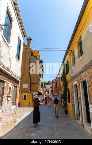 Touristes marchant dans une rue étroite colorée à Venise. Les gens se promènent dans une ruelle calme et ensoleillée à Venise, entourée de vieux bâtiments colorés et Banque D'Images