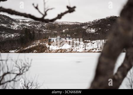 Un paysage hivernal serein dans les îles Lofoten, en Norvège, présente des collines enneigées et une cabane confortable nichée au bord d'un lac gelé, offrant un cadre paisible et rem Banque D'Images