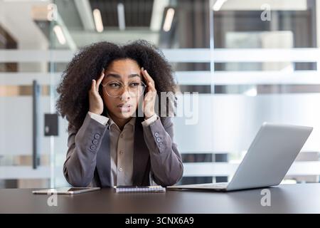 Jeune femme afro-américaine fatiguée et dépassée assise à son bureau dans le bureau et tenant sa tête dans ses mains. Banque D'Images