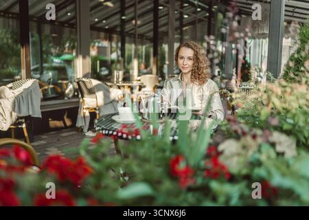 Une femme professionnelle est assise dans un café élégant, écrivant dans un cahier. La lumière du soleil coule à travers les grandes fenêtres, soulignant l'ambiance chaleureuse et elle Banque D'Images
