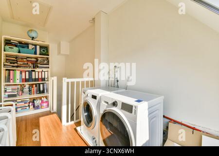 Une buanderie lumineuse et organisée avec des machines à laver modernes, des étagères de rangement remplies de livres, et un plancher en bois chaud qui améliore le sp Banque D'Images