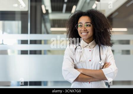 Photo d'une jeune femme afro-américaine médecin debout dans un bureau de clinique dans un uniforme blanc, les bras croisés sur sa poitrine et regardant sur le côté. Banque D'Images