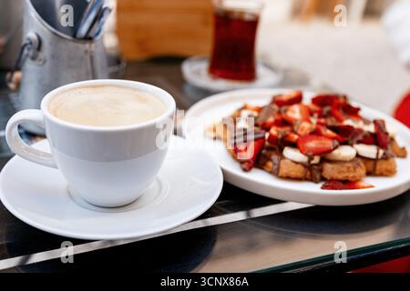 Une tasse de café à la vapeur accompagnée de délicieuses gaufres garnies de fraises fraîches et de sauce au chocolat, servies sur une table dans une atmosphère chaleureuse de café Banque D'Images