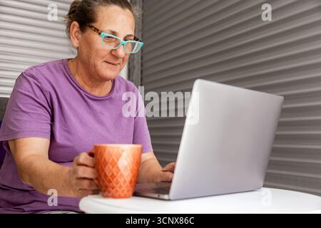 Femme senior avec des lunettes portant une chemise violette est confortablement assis à la maison, tenant une tasse tout en utilisant son ordinateur portable. Elle semble concentrée et satisfaite en lui Banque D'Images